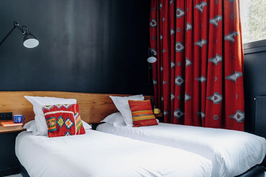 A double bed with white bedding and two pillows, a wooden headboard, a bedside table with a lamp, and red patterned curtains.
