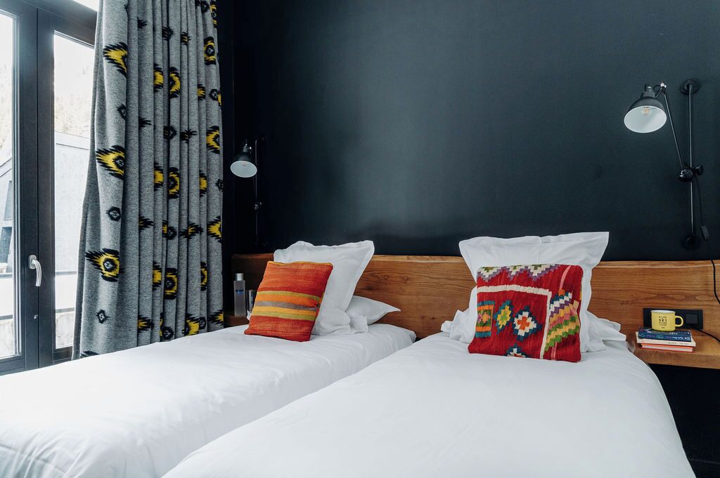 A hotel room with two beds, grey curtains with a pattern, a wooden headboard, white bedding, a red and yellow pillow, a small table with a mug, and a wall-mounted lamp.