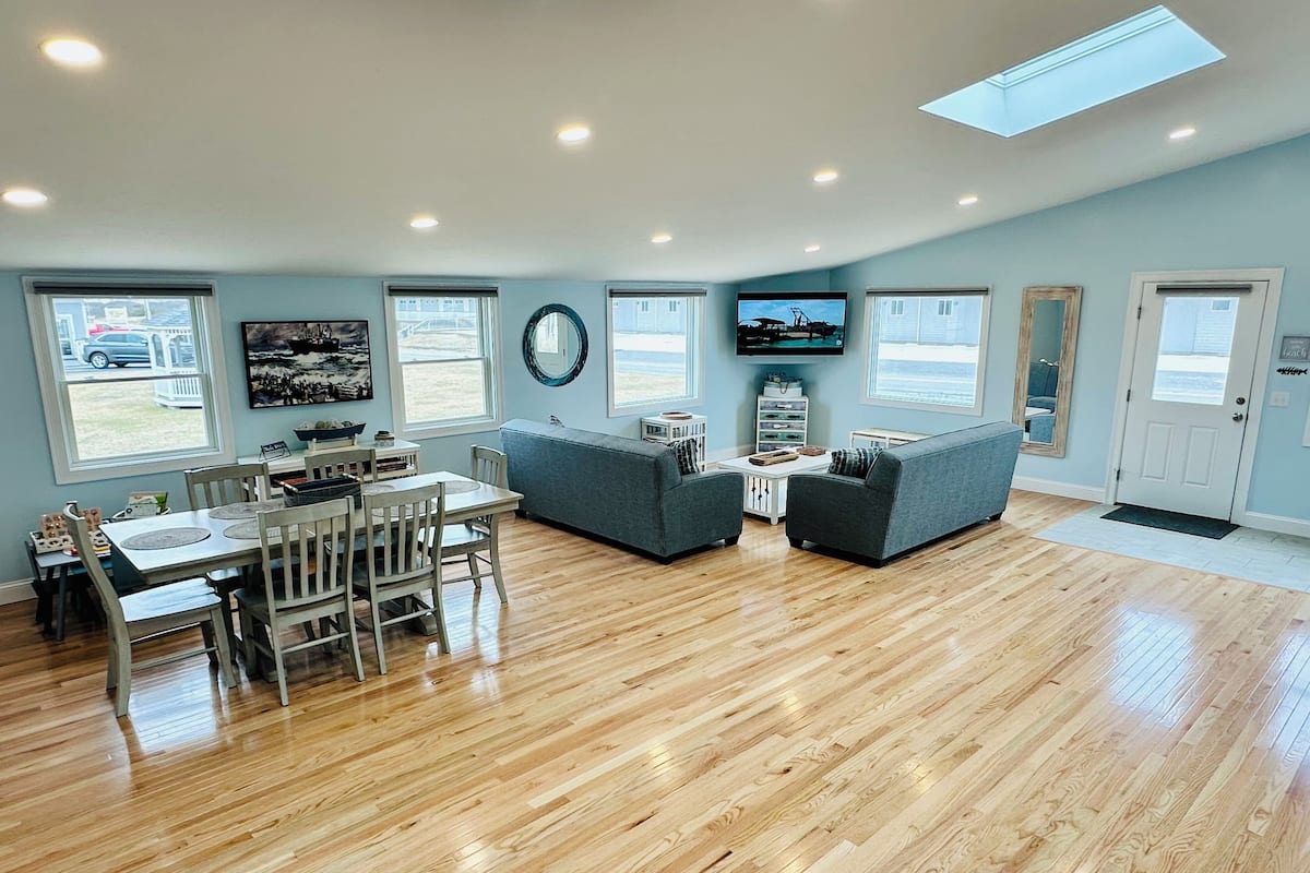 A spacious living room with a wooden floor, a dining area with a table and chairs, a gray sofa, and a television mounted on the wall.