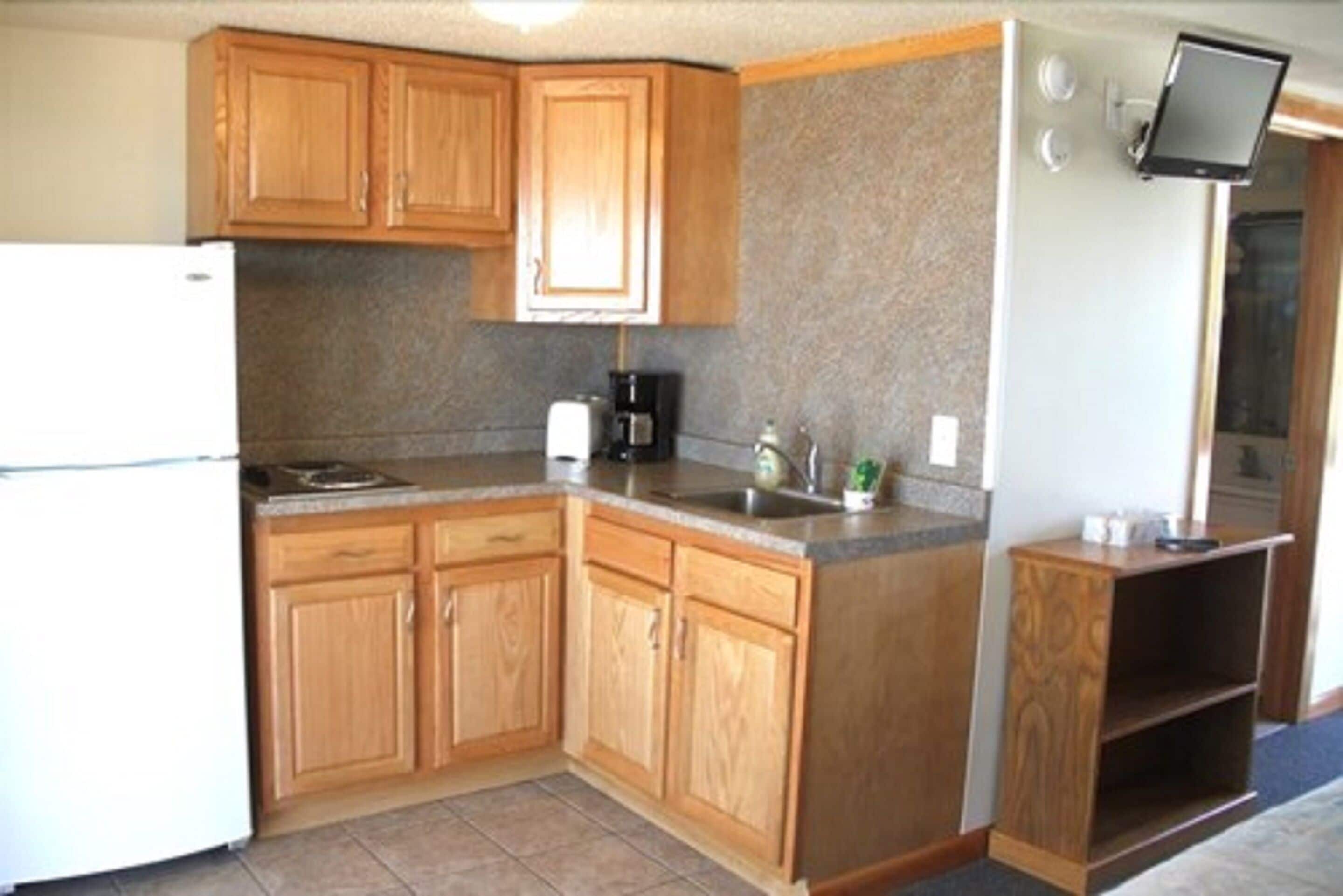 A kitchen with wooden cabinets, a refrigerator, a coffee maker, and a flat-screen TV mounted above the counter.