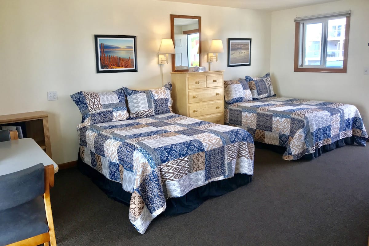 A bedroom with two beds, a dresser, a desk, and two windows.