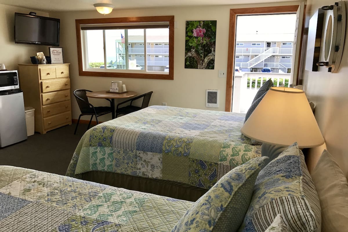 A hotel room with two beds, a desk, a chair, a TV, a microwave, a dresser, and a window with a view of a building and a balcony.