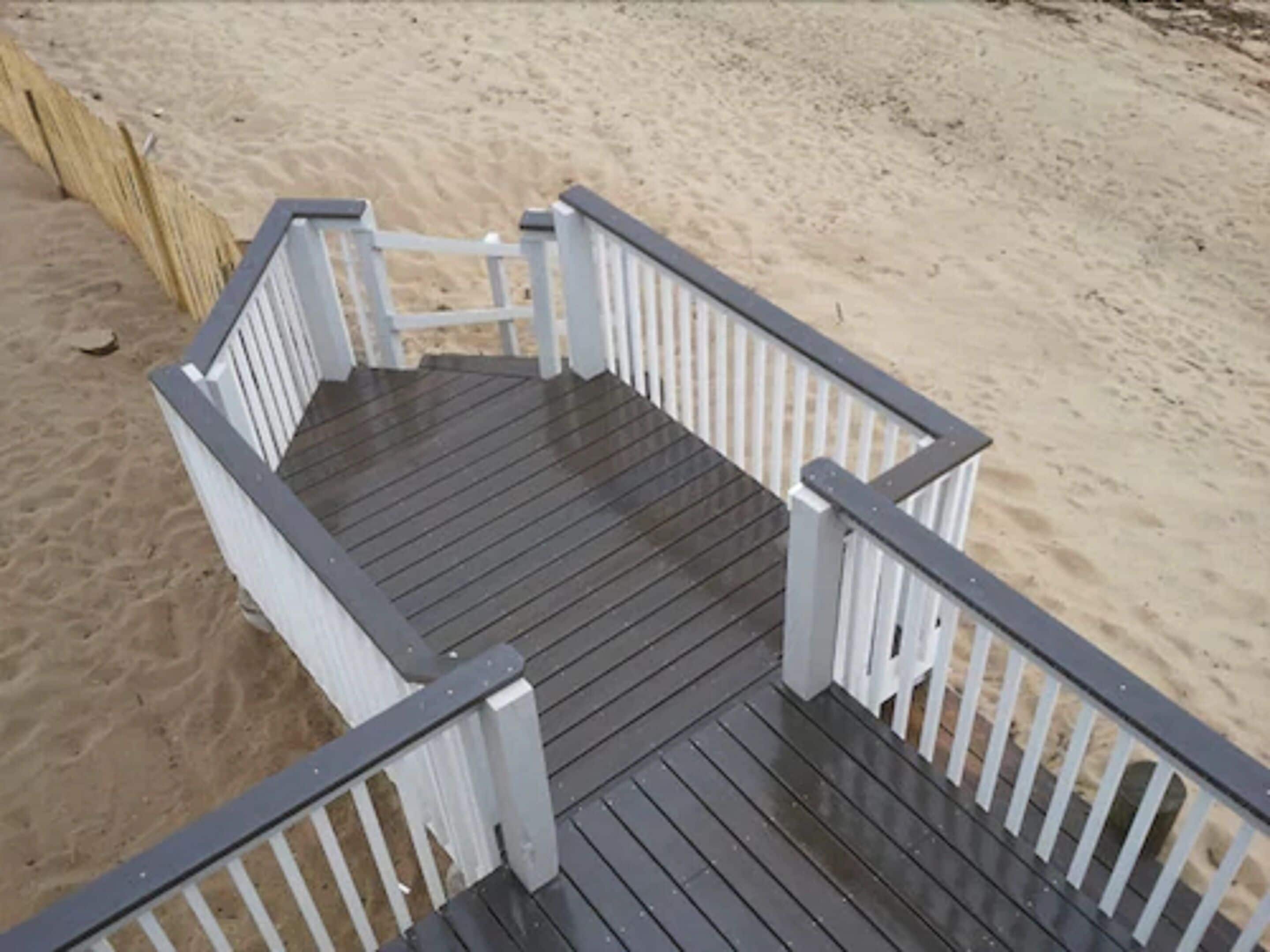 Um deck de madeira com guarda-corpos brancos que dá para uma praia de areia.