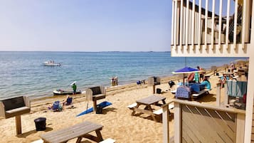Relax on the beach with picnic tables, lounge chairs, and a barbecue setup, perfect for dining and outdoor activities.