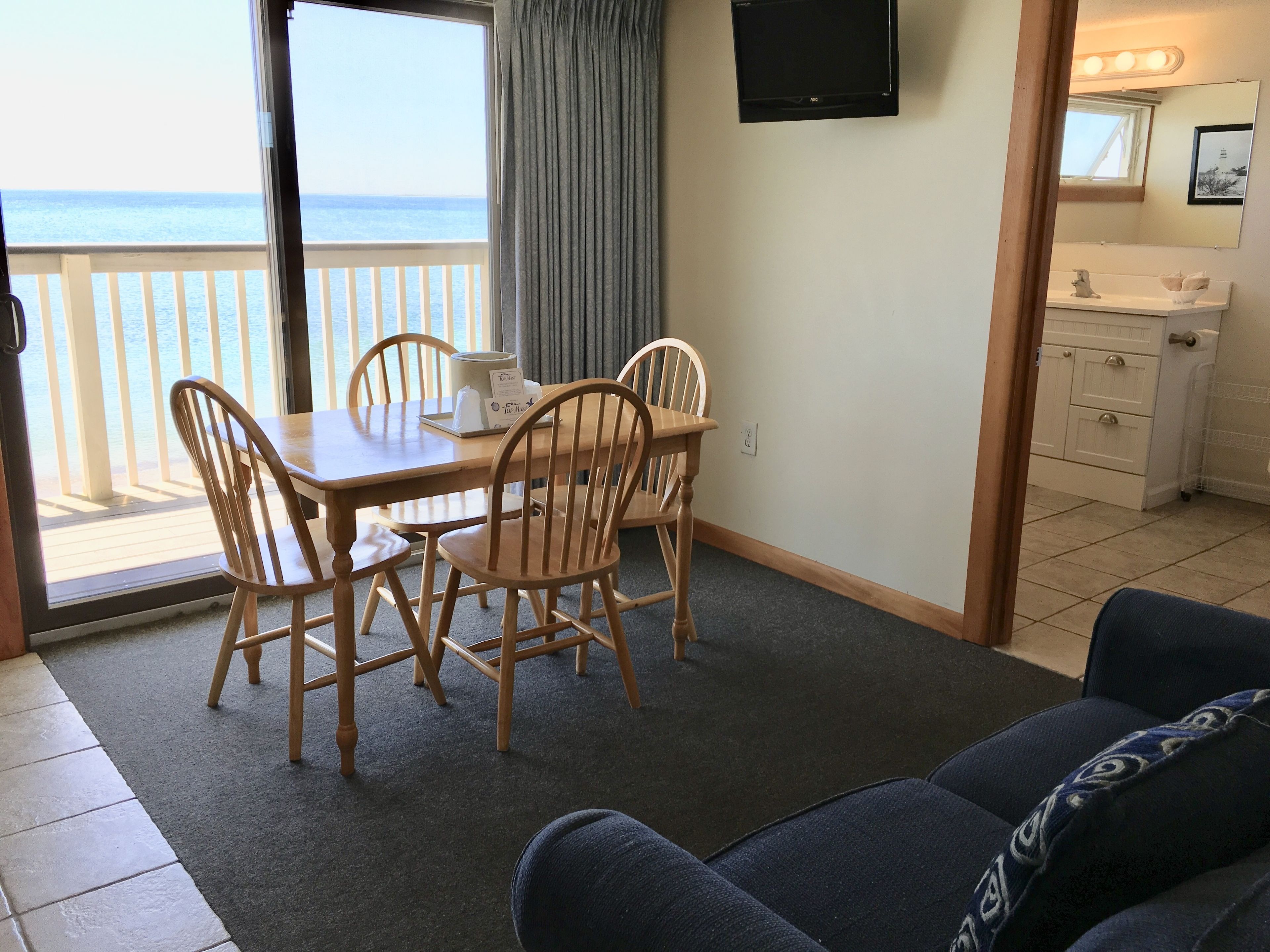 A room with a dining table and chairs, a TV mounted on the wall, a view of the ocean through a sliding door, and a small kitchenette.