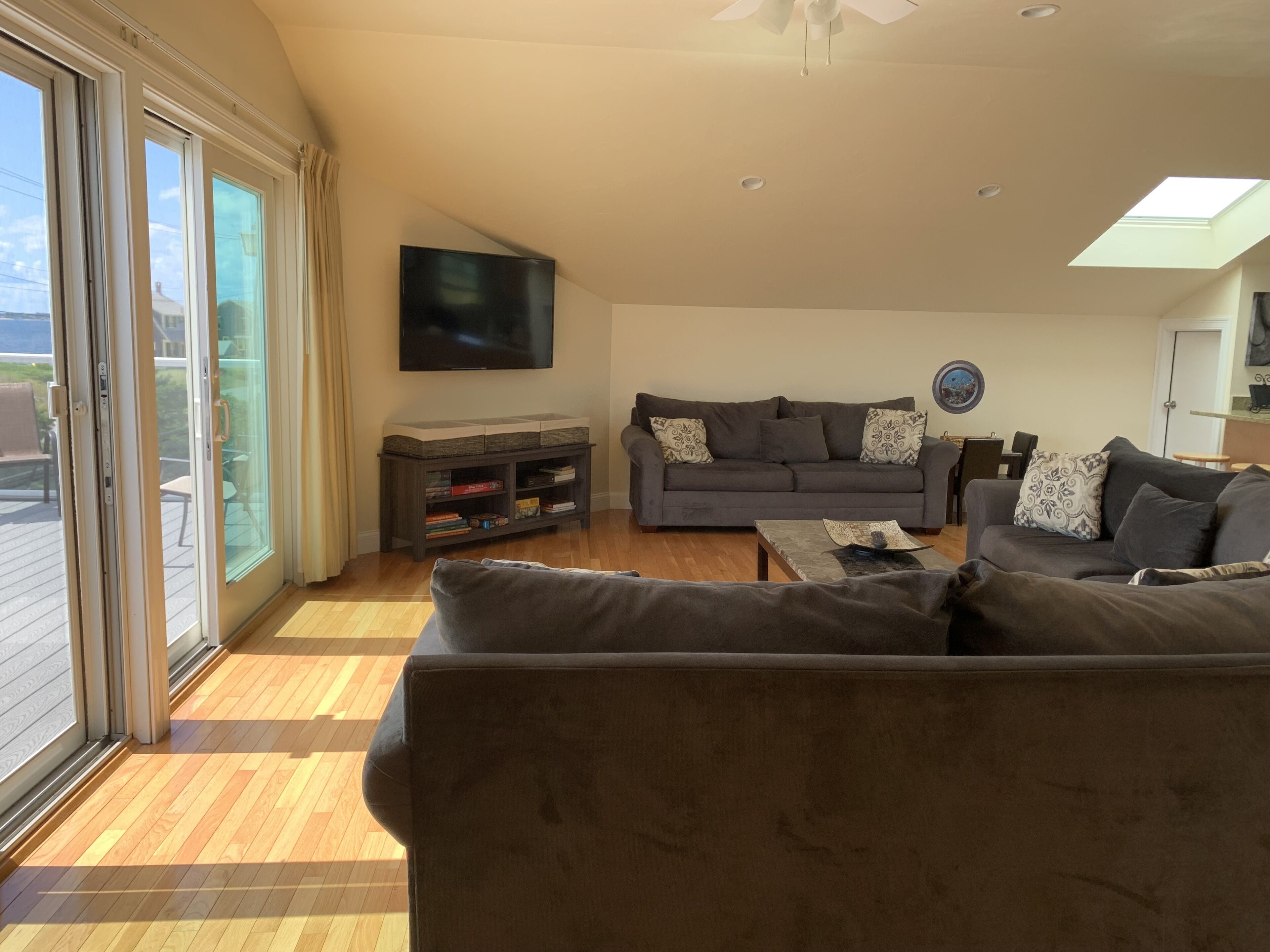 A living room with a grey sofa, a coffee table, and a TV mounted on the wall. There is a sliding glass door leading to an outdoor area.