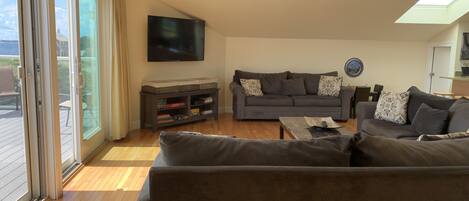 A living room with a grey sofa, a coffee table, and a TV mounted on the wall. There is a sliding glass door leading to an outdoor area.