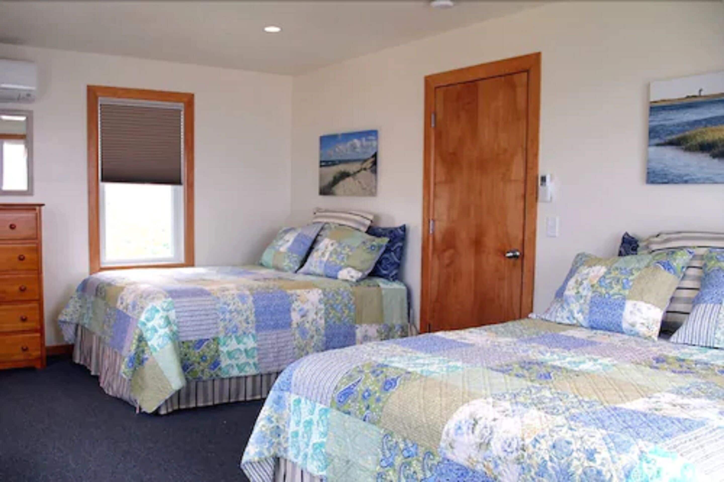 A bedroom with two beds, a wooden door, a window with blinds, a dresser, and a painting on the wall.