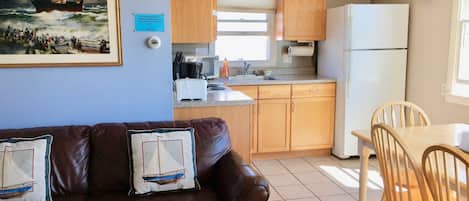 A compact kitchen with wooden cabinets, a dining table with chairs, a refrigerator, and a painting on the wall.