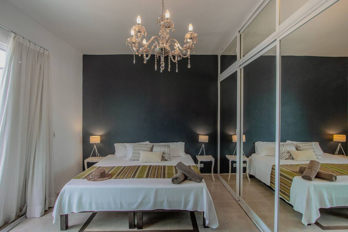 Ein Hotelzimmer mit zwei Betten, einer Chandelüster, Nachttischlampen, einem Spiegel und einem Fenster mit Vorhängen.