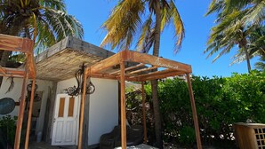 Un lit en bois, surmonté d’un dais, installé dans un espace extérieur sablonneux, entouré de palmiers et d’un petit bâtiment doté d’une porte.