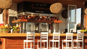 Un bar de plage avec des chaises blanches, un panier de fruits et diverses bouteilles exposées sur le comptoir.