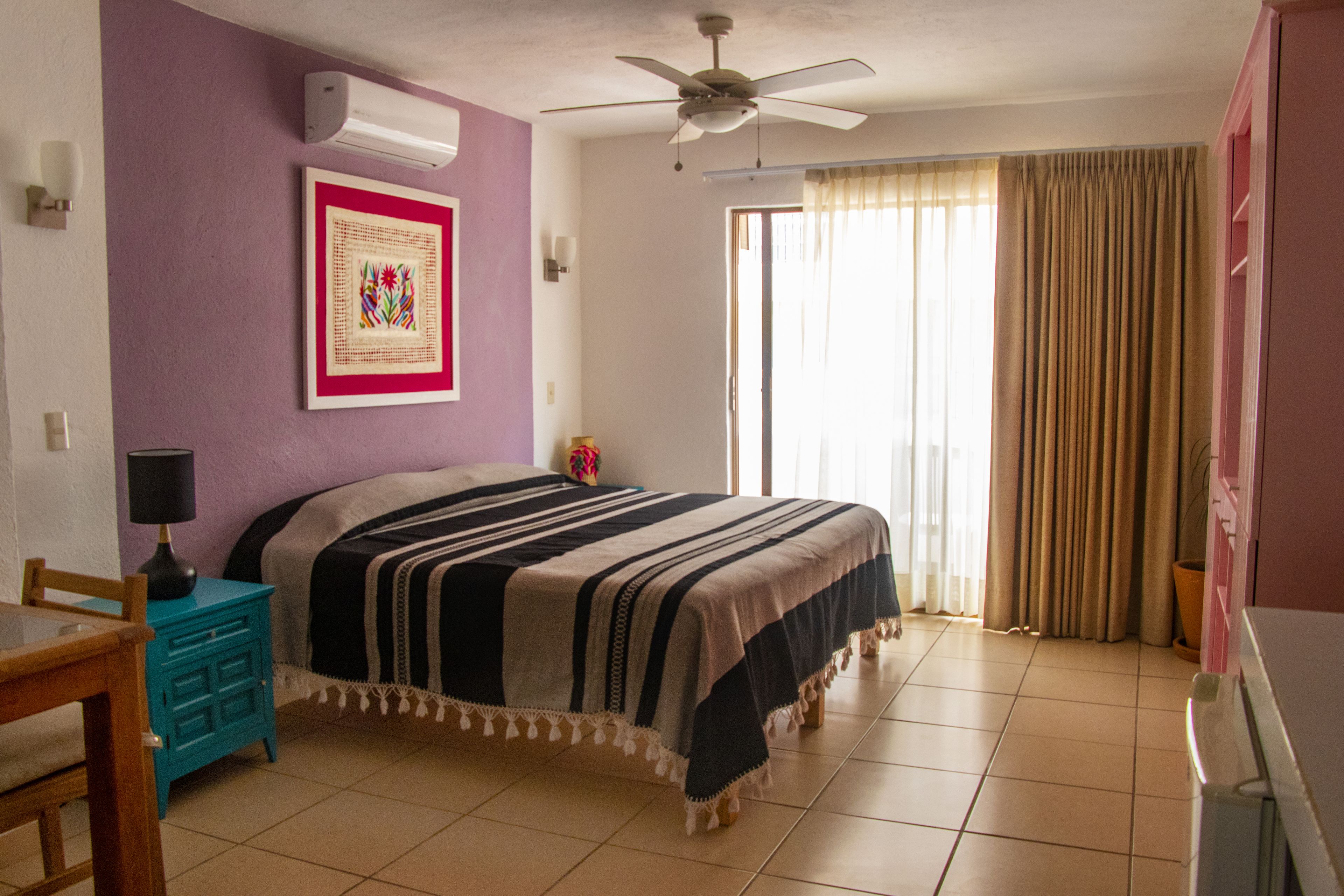Un dormitorio con cama, ventilador de techo, ventana con cortinas, una mesita con lámpara y un cuadro en la pared.