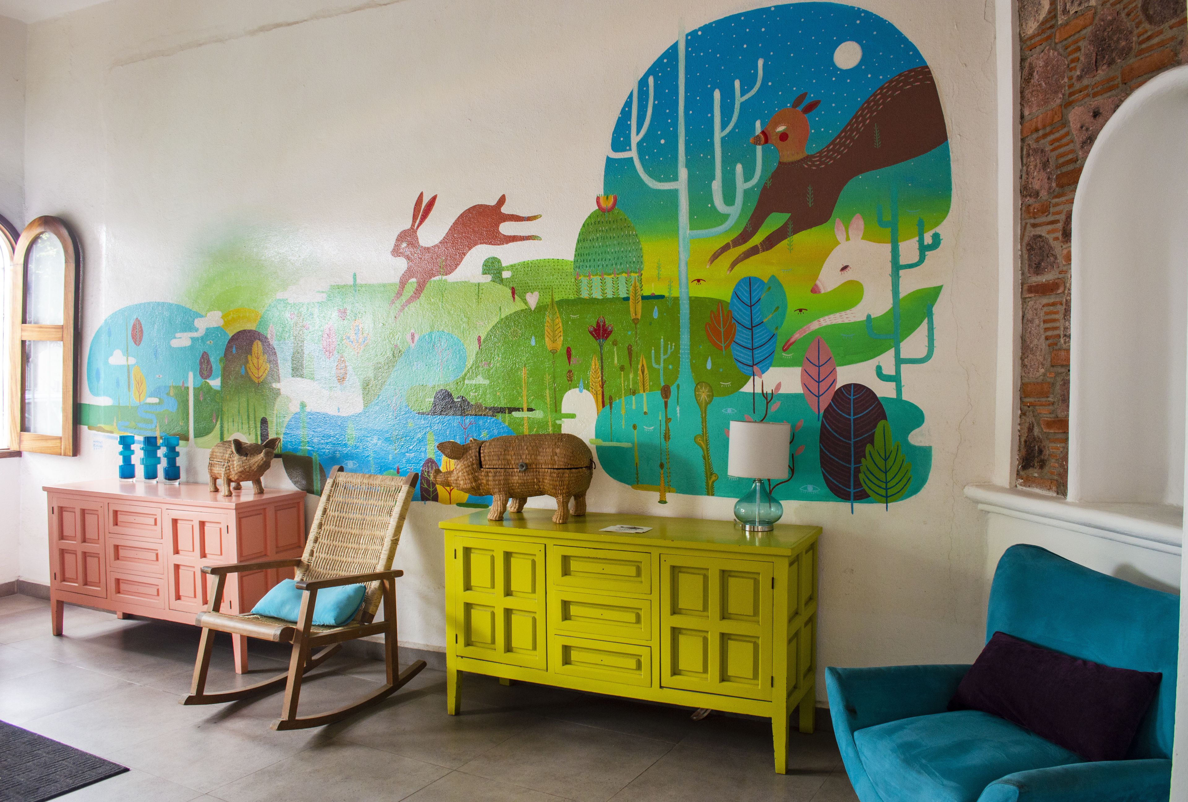 A room with a colorful mural, a rocking chair, a yellow cabinet, and a blue sofa.