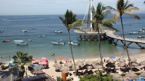 Een strand met rieten parasols, palmbomen en boten op het water.