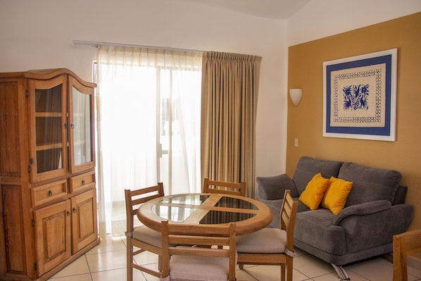 A living room with a round glass table, wooden chairs, a grey sofa with yellow cushions, and a wooden cabinet.