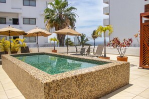 A rectangular pool with mosaic tiles, surrounded by lounge chairs and umbrellas, with a view of palm trees and a building in the background.