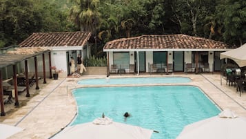 Piscina com dois guarda-sóis, rodeada por espreguiçadeiras e mesas, com um edifício ao fundo.