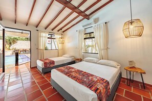 A hotel room with two beds, a small table, a hanging light fixture, and a view of an outdoor pool area. - Hotel Quimbaya (San Jerónimo)