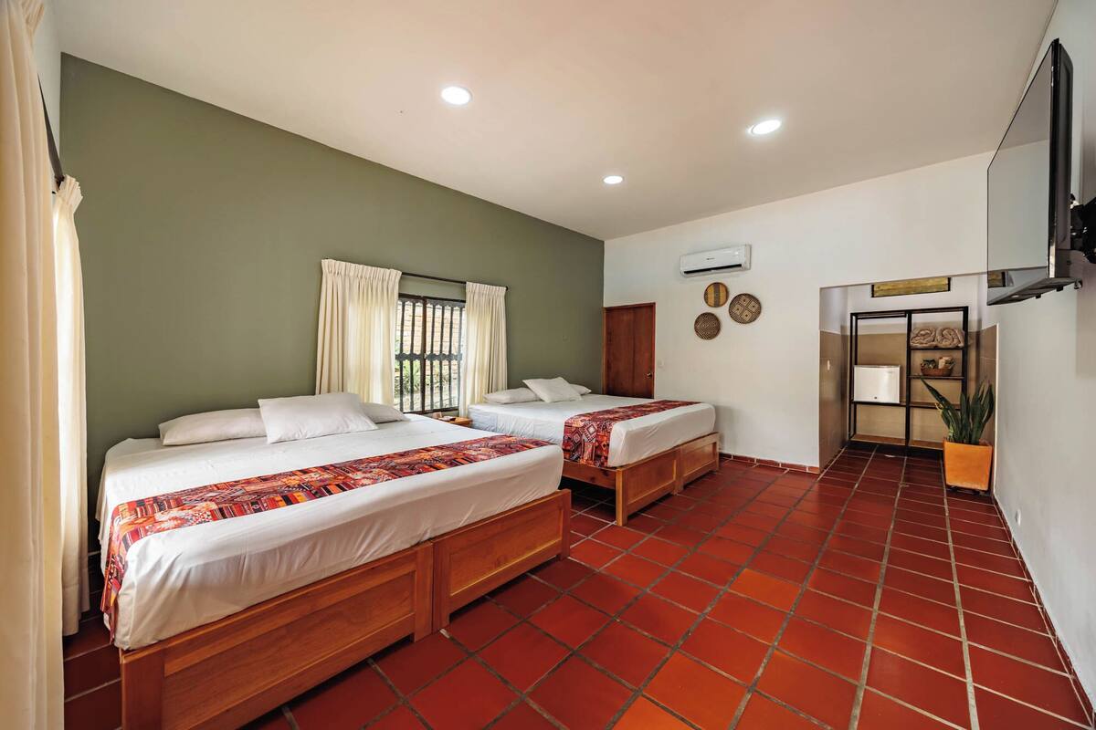 A hotel room with two beds, a window with curtains, a wall-mounted air conditioner, and a TV.