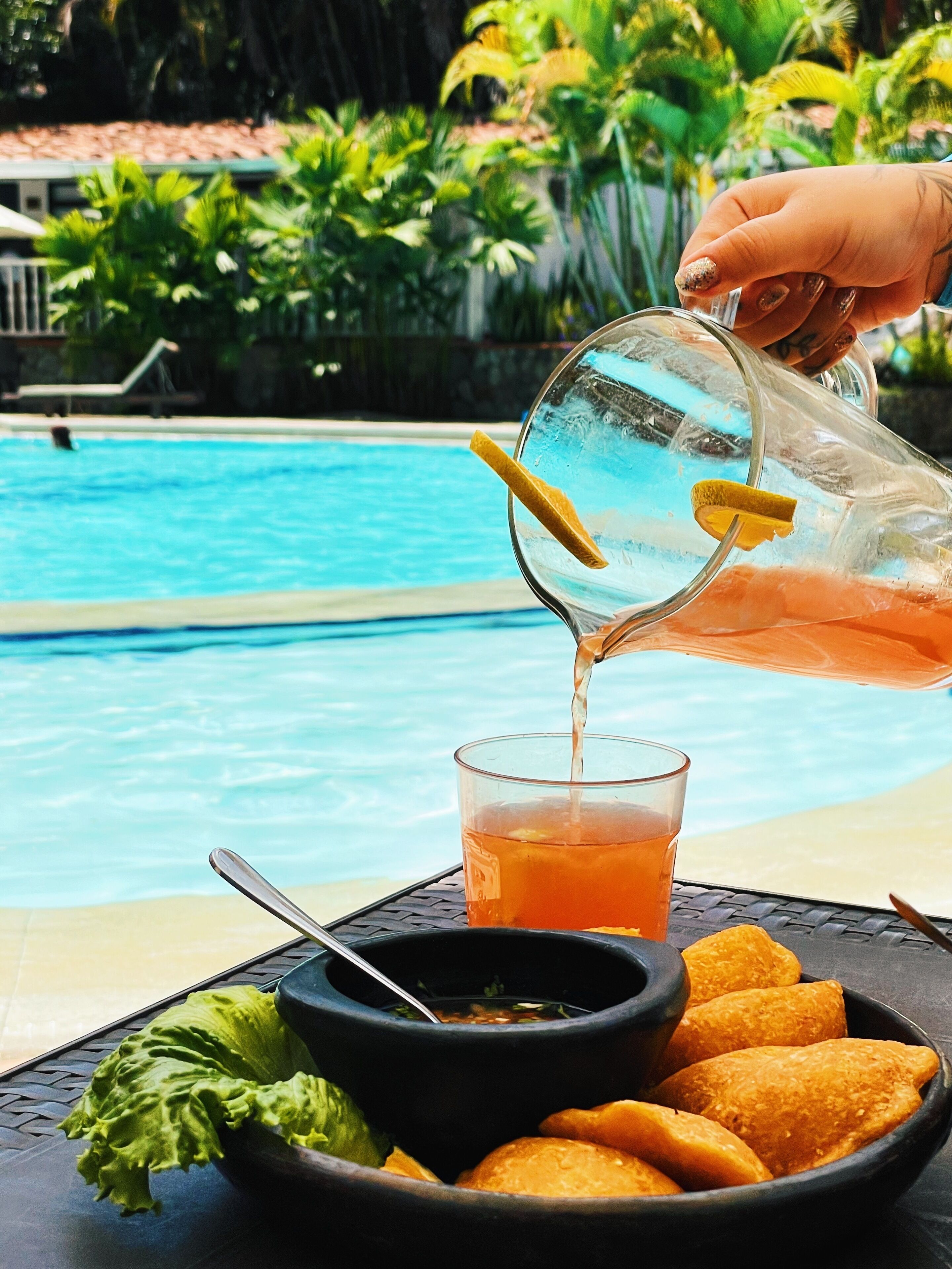 A hand pouring a drink into a glass near a poolside table with a bowl of food and a spoon.