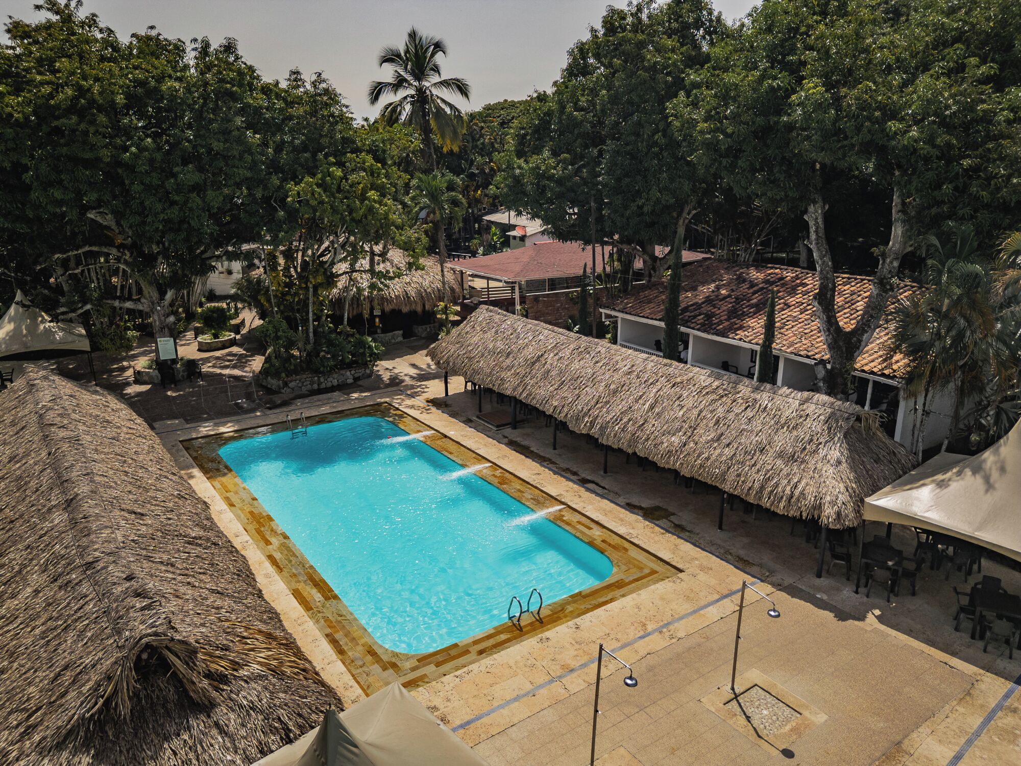 Un área de piscina con una estructura de techo de paja, rodeada de árboles y un edificio con tejado de azulejos.