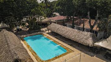 Un área de piscina con una estructura de techo de paja, rodeada de árboles y un edificio con tejado de azulejos.