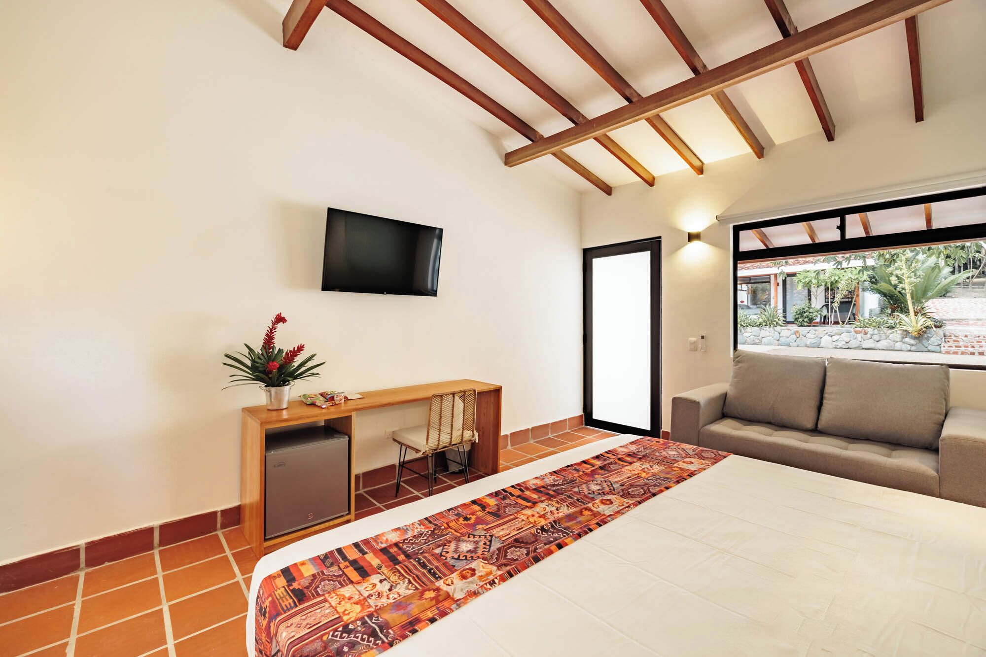 Una habitación de hotel con una cama, un televisor montado en la pared, una pequeña mesita de madera con una planta, un sofá y una alfombra estampada.