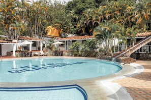 A swimming pool with a tropical setting, surrounded by palm trees and a covered area with outdoor seating. - Hotel Quimbaya (San Jerónimo)