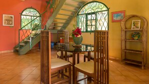 A dining area with a glass table, wooden chairs, a vase with flowers, a staircase with green railings, and a wall with a picture frame. - Sundown Villa -Negril (Negril)