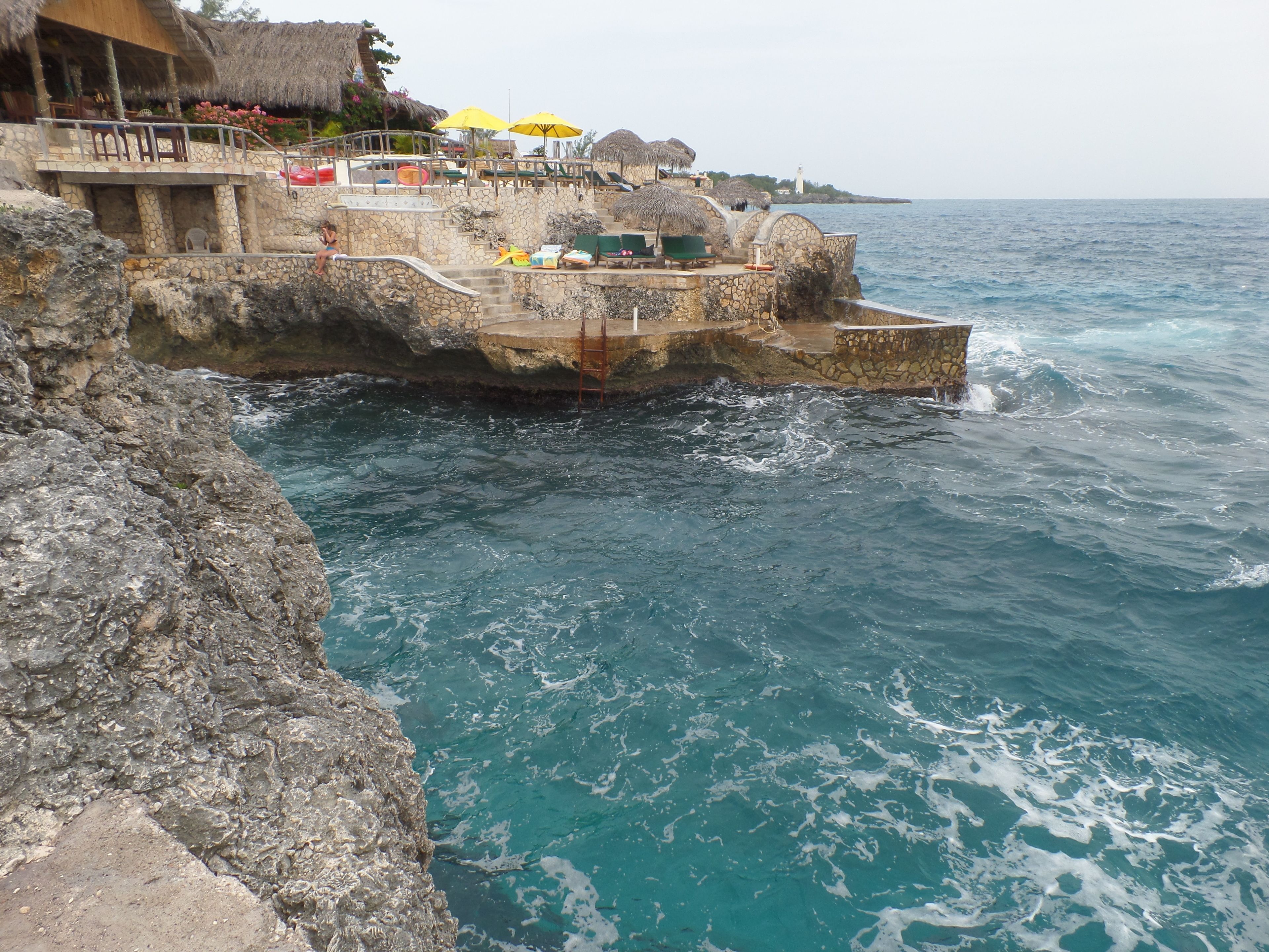 A coastal stone structure with a pool and umbrellas, overlooking the sea.