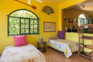 Una habitación con dos camas, un ventilador, una ventana con rejas y una silla.