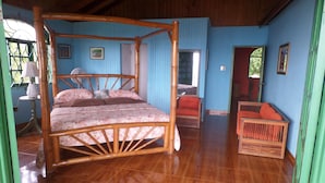 A four-poster bed with a floral bedspread, a wooden floor, a red sofa, and a fan in a room with blue walls and a wooden ceiling. - Sundown Villa -Negril (Negril)