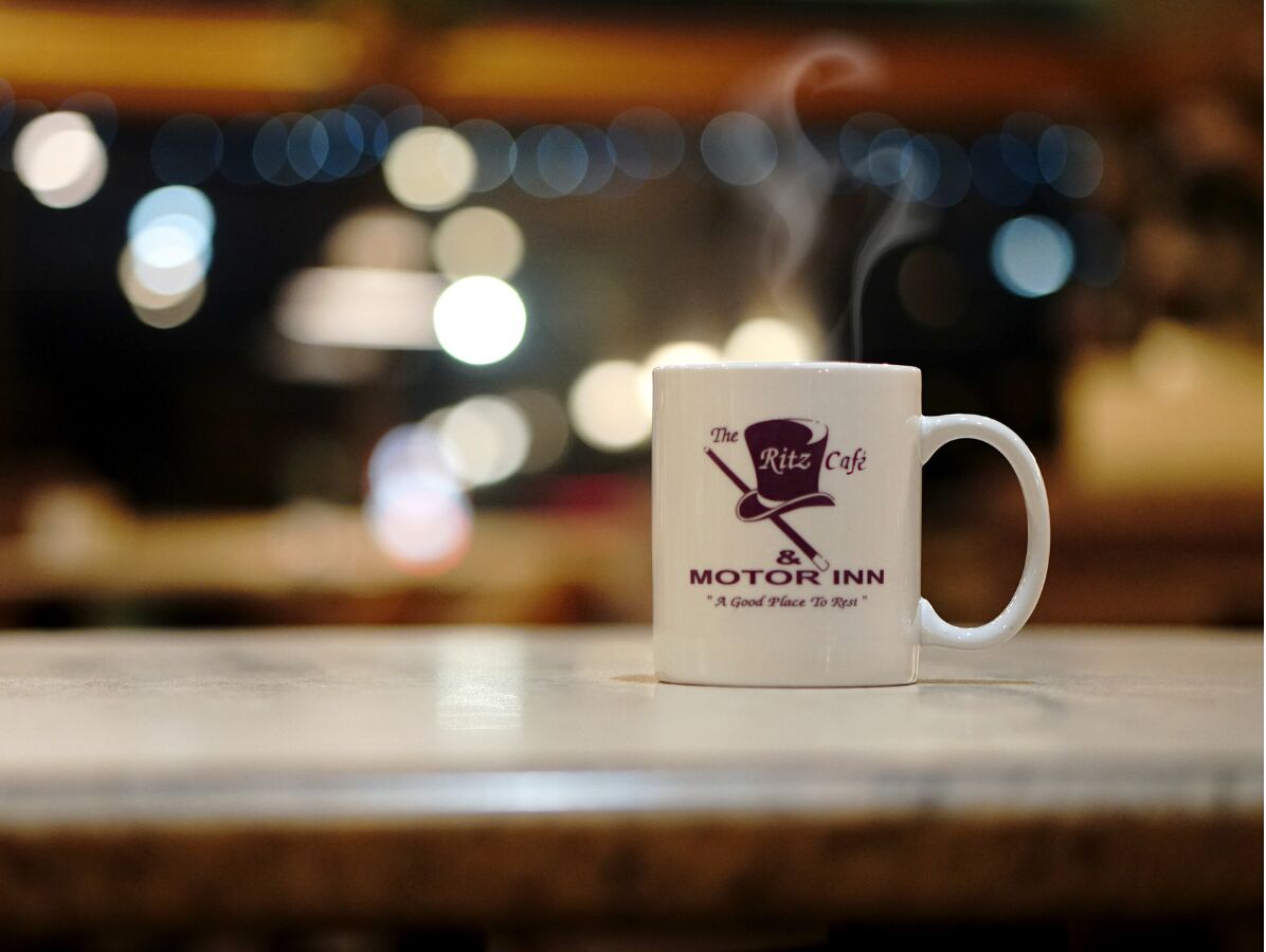 A white mug with "The Ritz Cafe & Motor Inn" logo and slogan on a table.