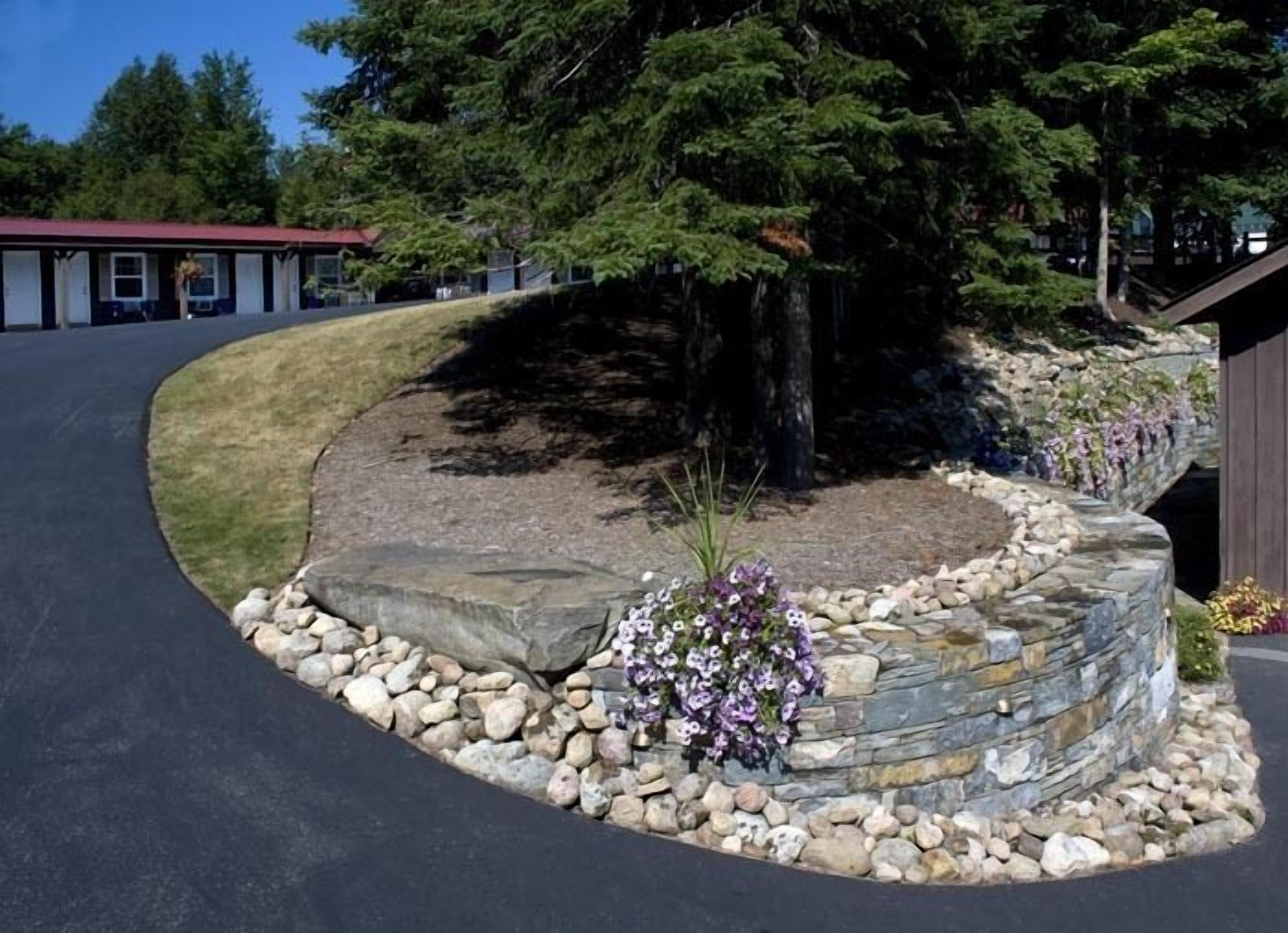 A landscaped area with a stone retaining wall, a large tree, and a building in the background.