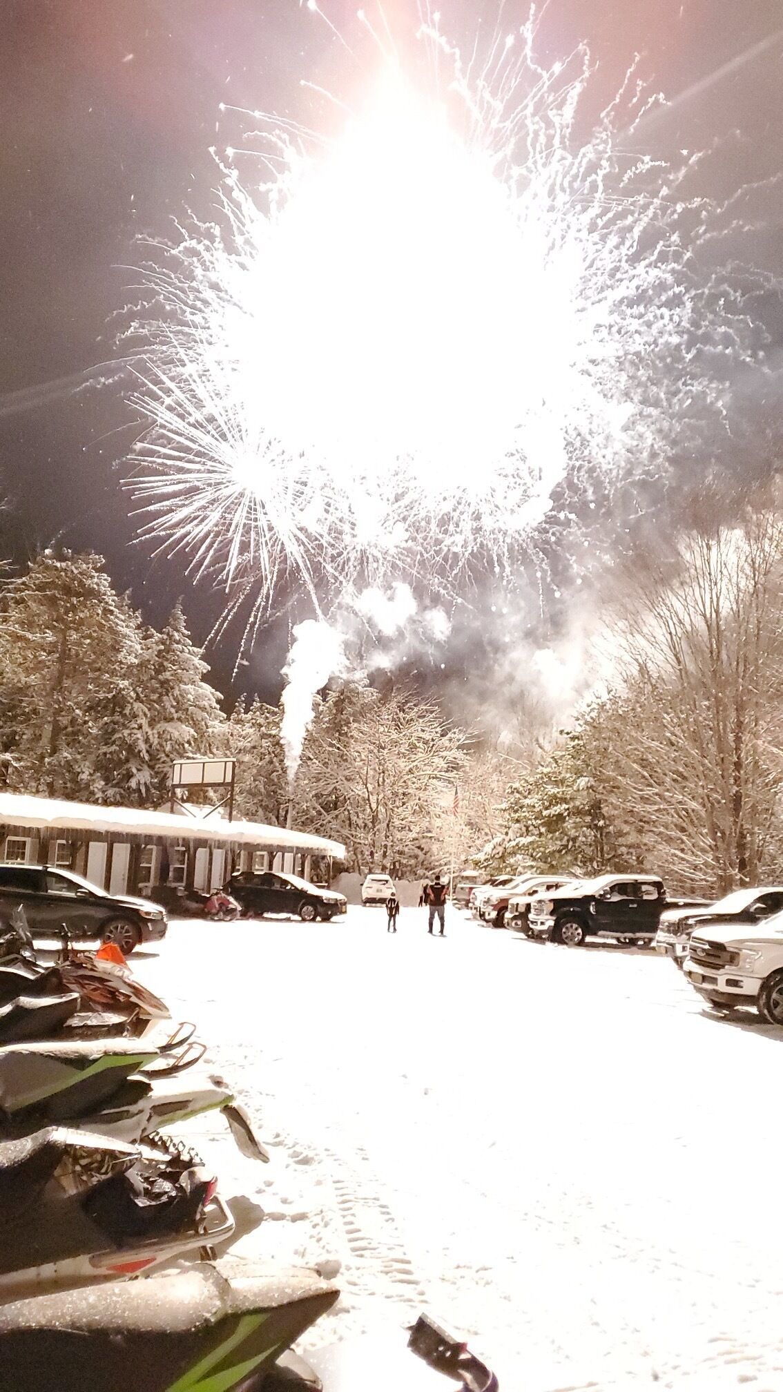Un parking rempli de véhicules, avec un feu d’artifice en arrière-plan.
