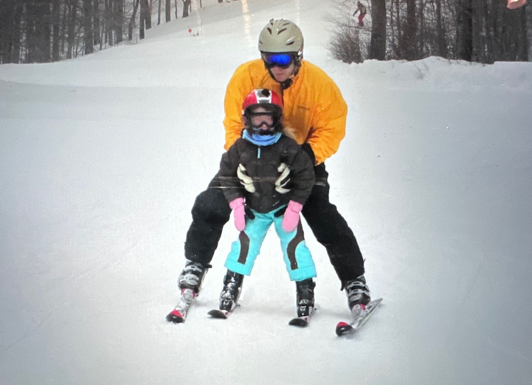 Een besneeuwde helling met een persoon in een oranje jas en helm, en een kind in ski-uitrusting zwart en roze.