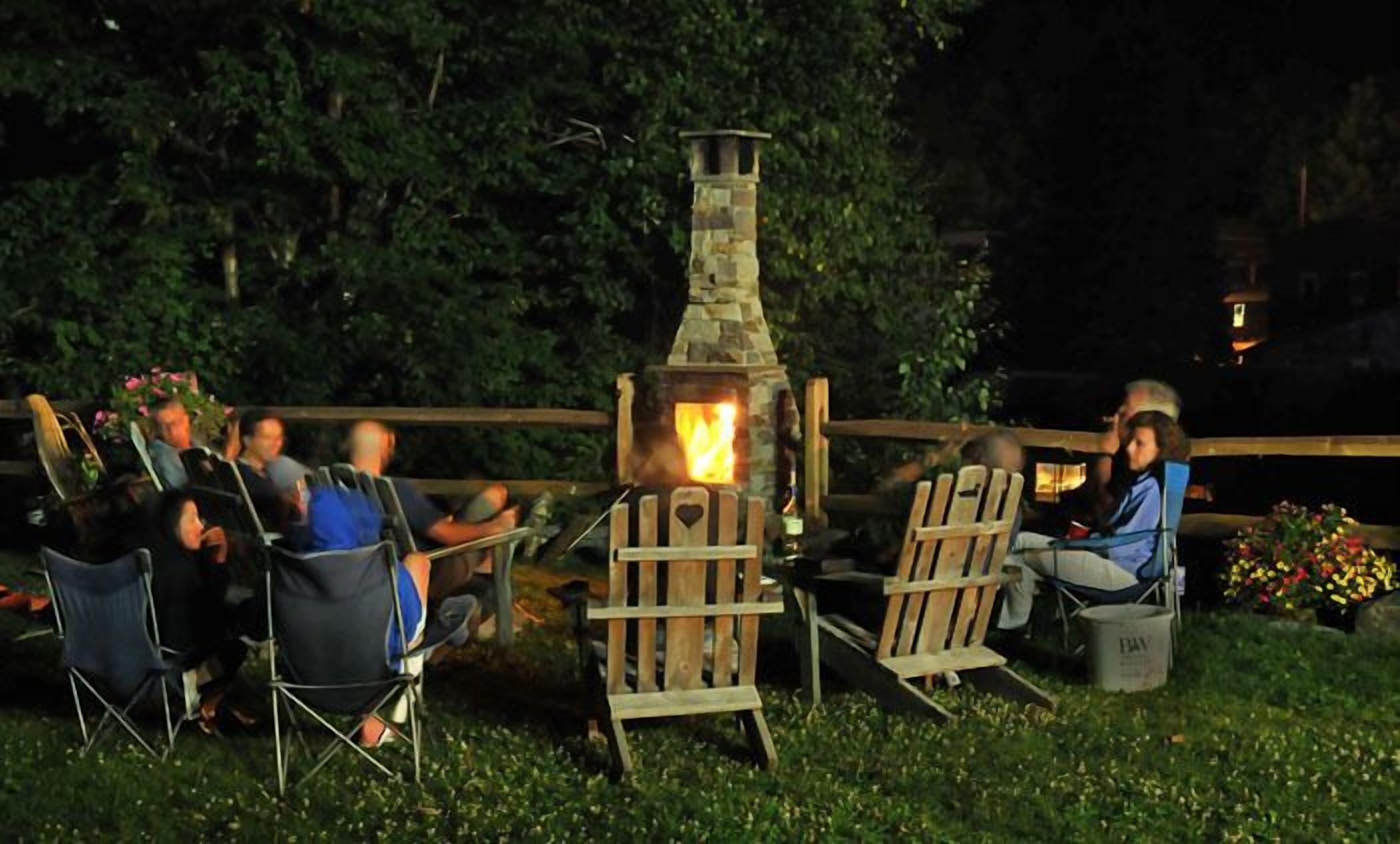 A group of people seated around a stone-built outdoor fireplace in a garden setting.