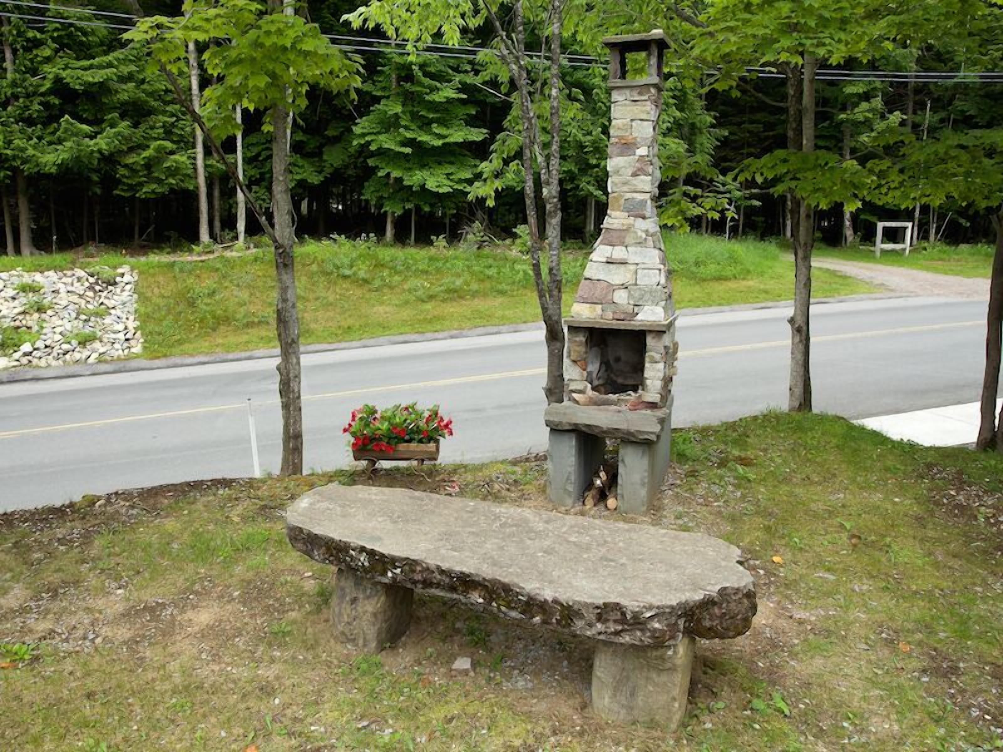 A stone-built outdoor fireplace with a bench in front, surrounded by trees and a road.