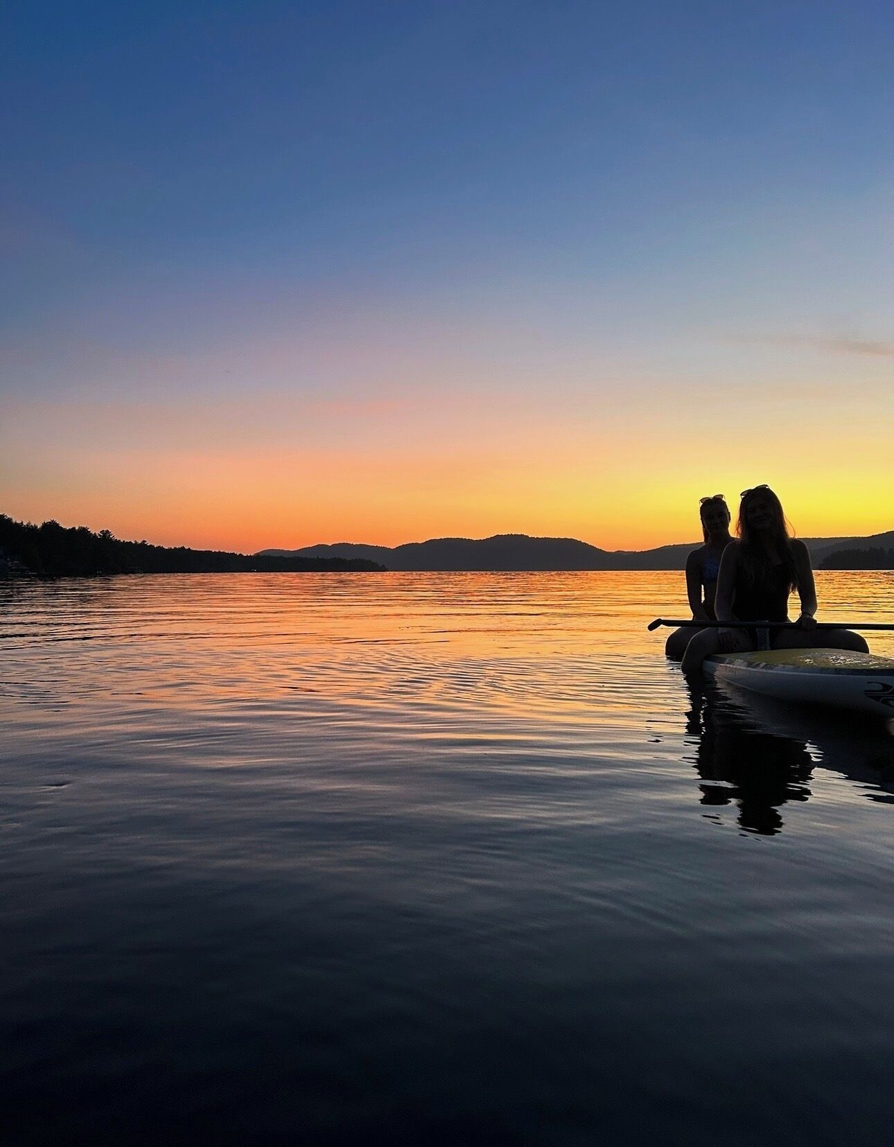 Two people on a paddleboard in calm water with a sunset in the background.