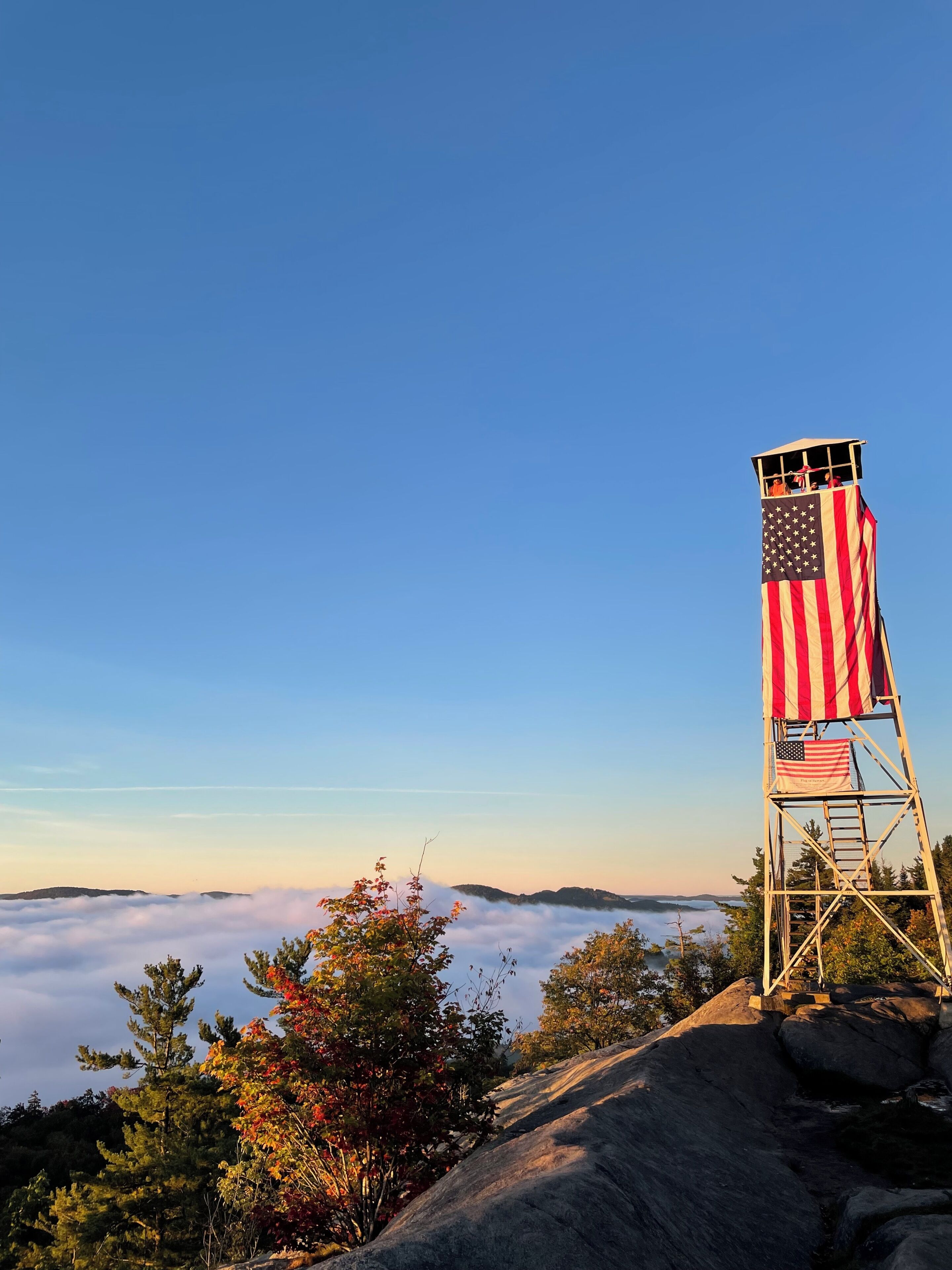 Uma torre de observação com uma bandeira americana, rodeada por árvores e um céu claro.