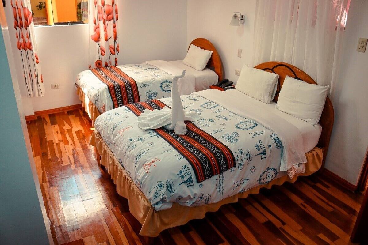 A hotel room with two beds, each with a wooden headboard, white bedding, and a decorative swan pillow. The room has wooden flooring and white curtains with a floral pattern.
