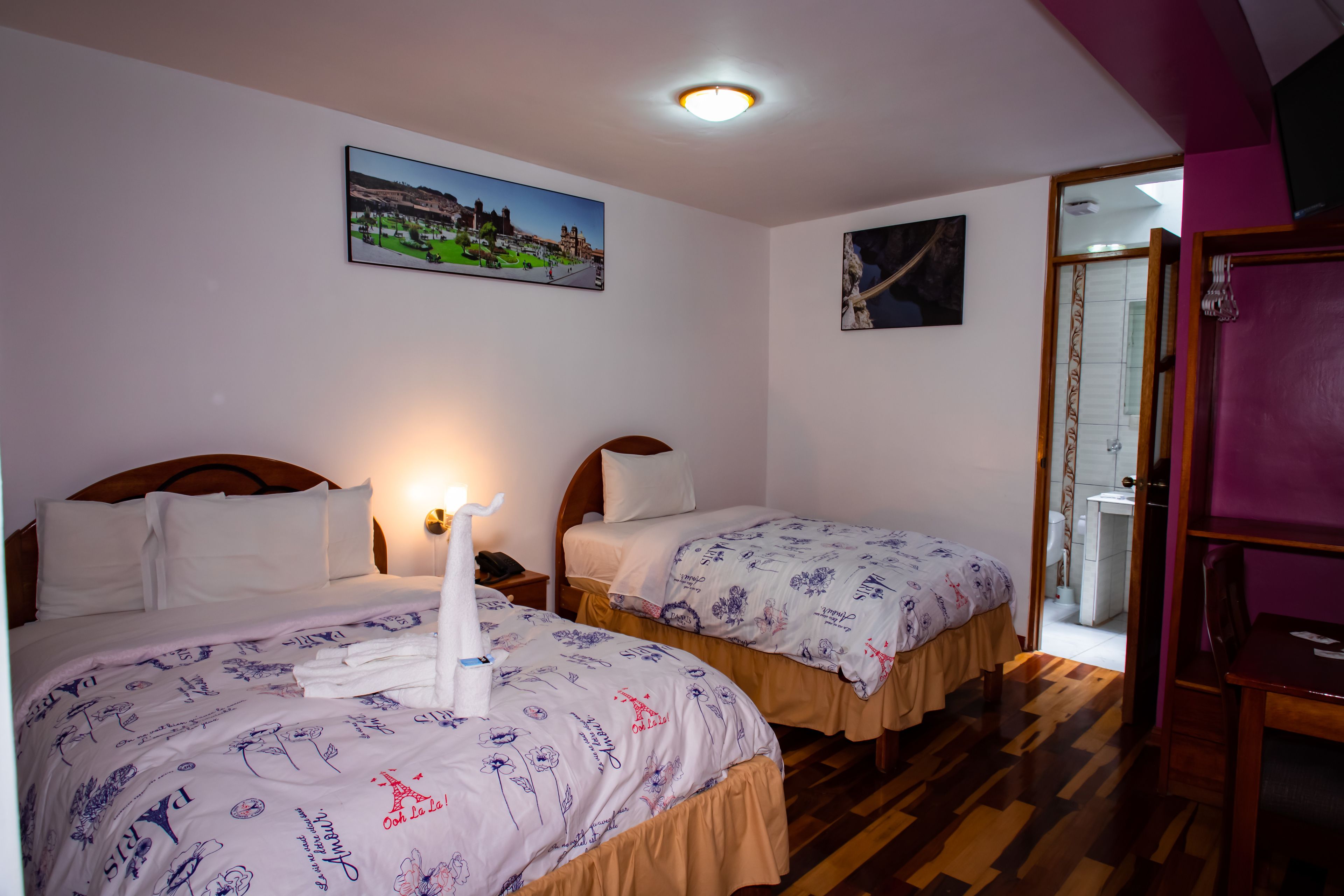 Habitación de hotel con dos camas, suelo de madera, un cuadro de un paisaje urbano y un baño visible a través de una puerta abierta.