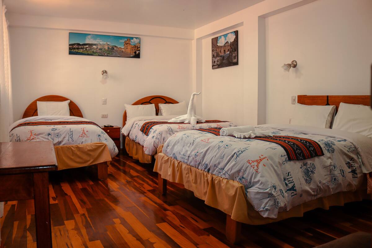 A hotel room with three single beds, wooden flooring, and two paintings on the walls.