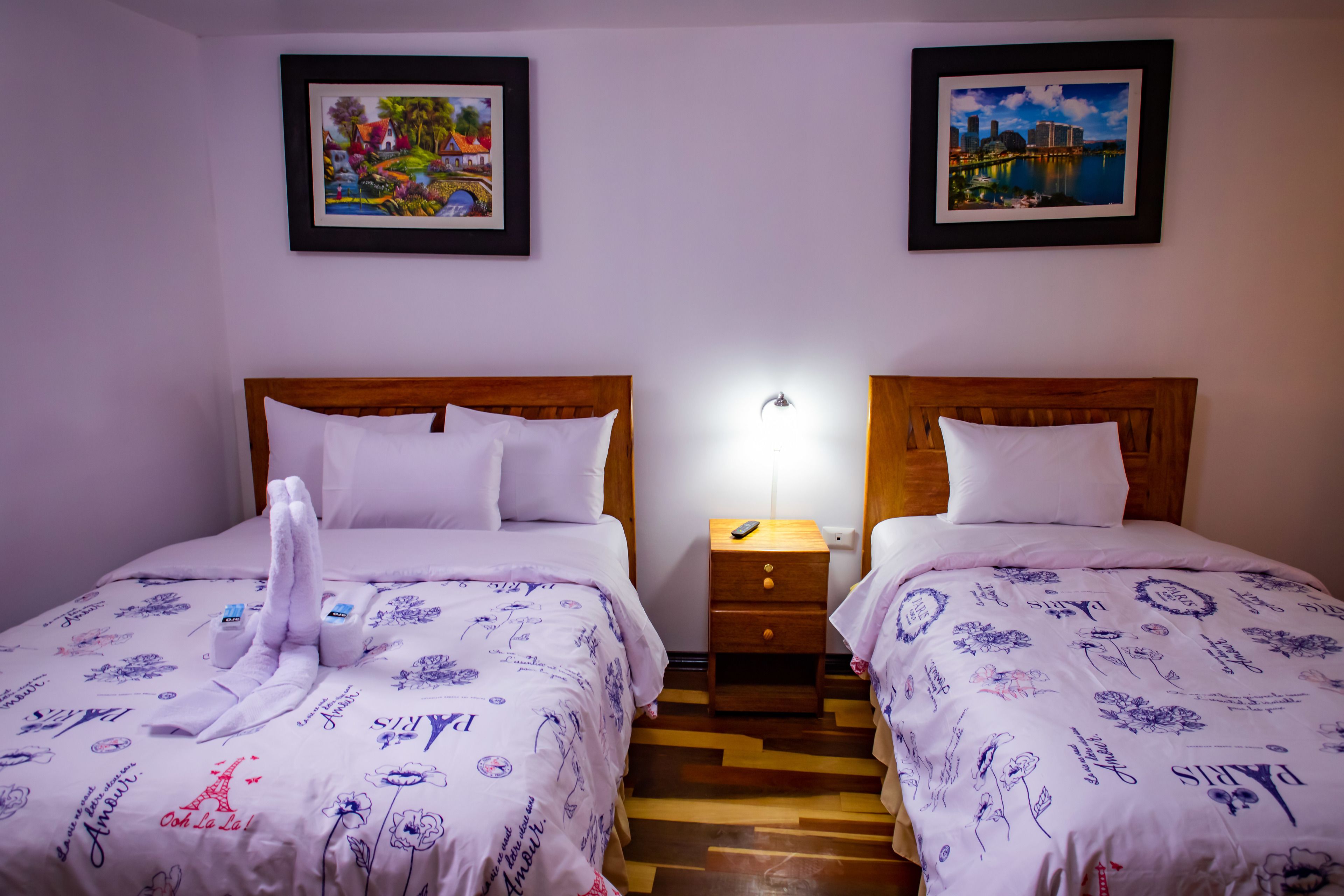A hotel room with two beds, each with a wooden headboard, white bedding, and a small bedside table with a lamp. There are two framed pictures on the wall above the beds.