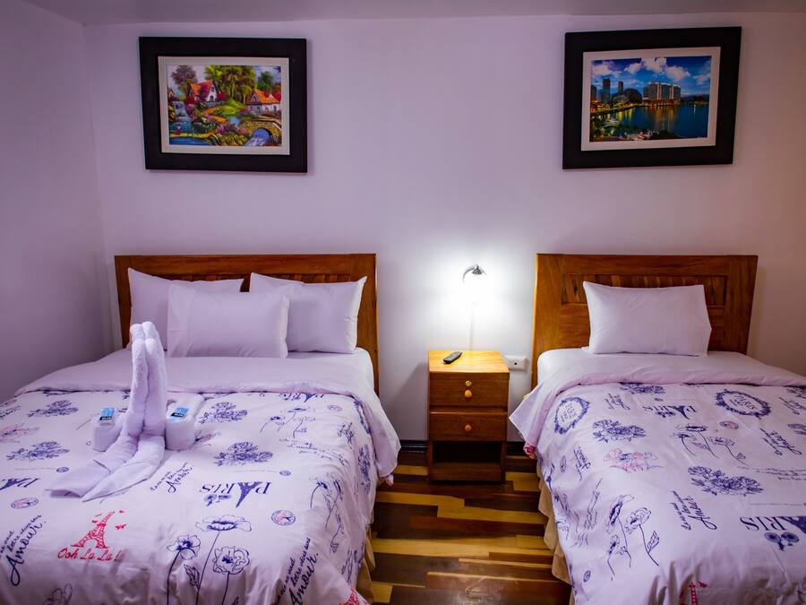 Een hotelkamer met twee bedden, elk met een houten hoofdbord, wit beddengoed en een klein nachtkastje met een lamp. Boven de bedden hangen twee ingelijste foto's.