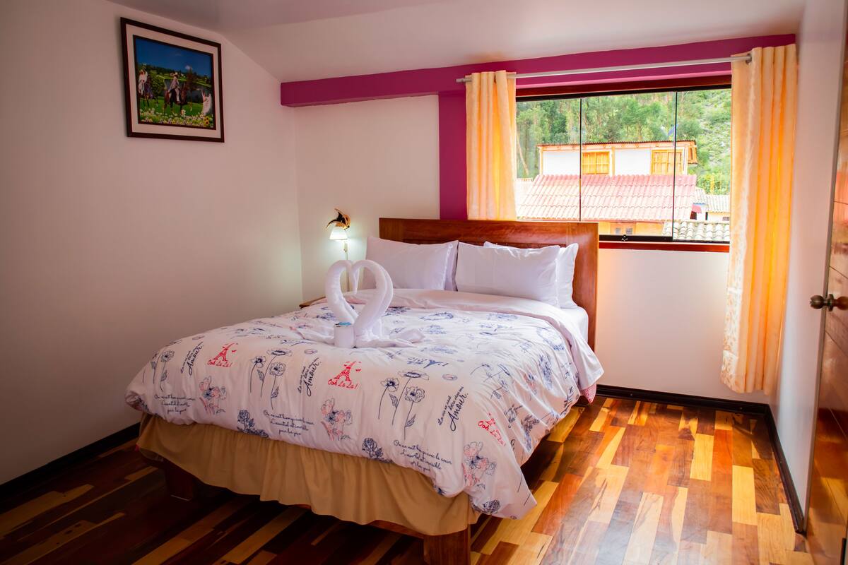 A hotel room with a bed, a window with curtains, a wooden floor, and a wall-mounted lamp.