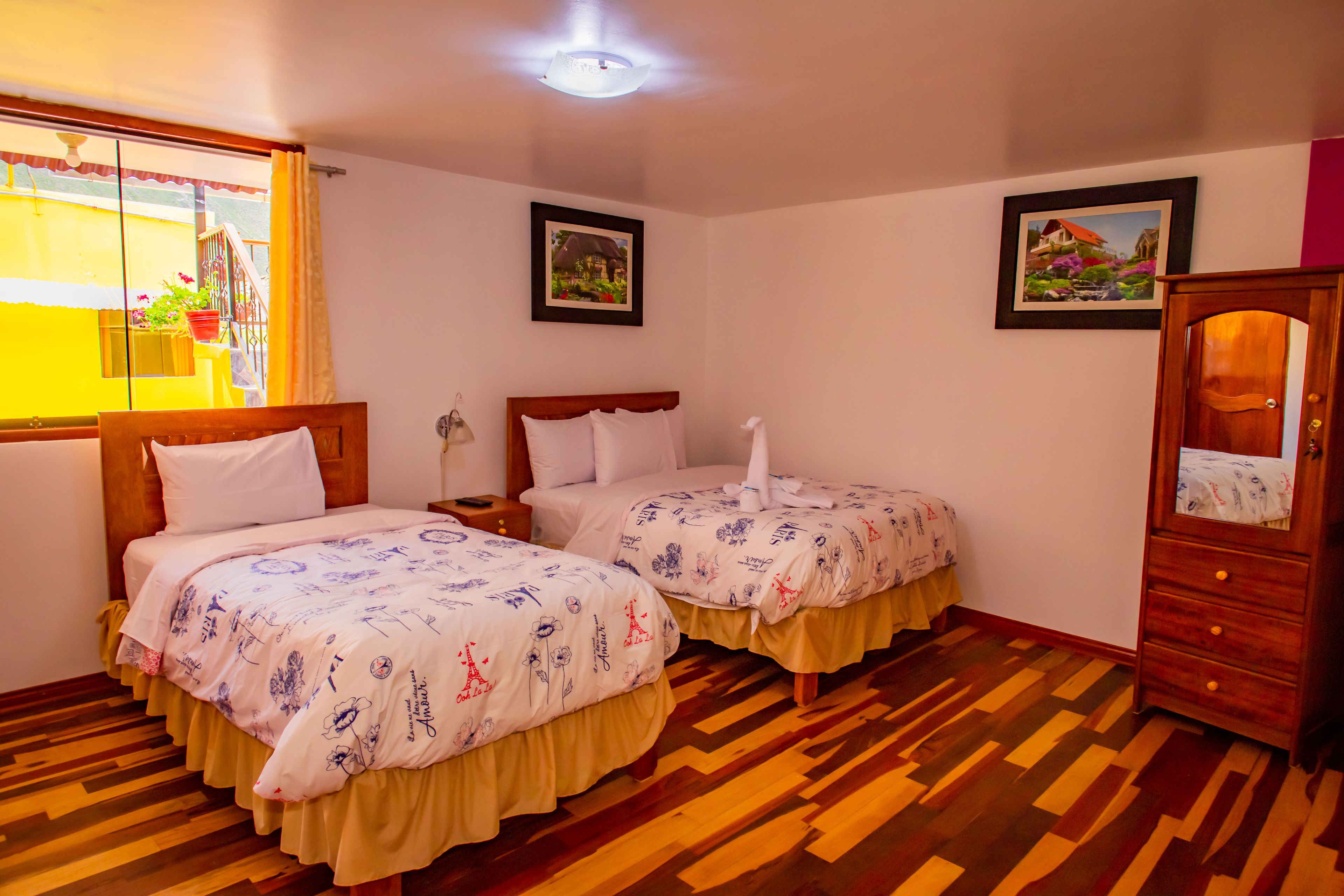 A hotel room with two beds, wooden flooring, a wooden wardrobe, and two framed pictures on the walls.