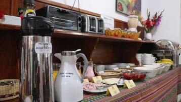 Disposição de buffet com dispensadores de água e leite quentes, uma torradeira e diversos alimentos.