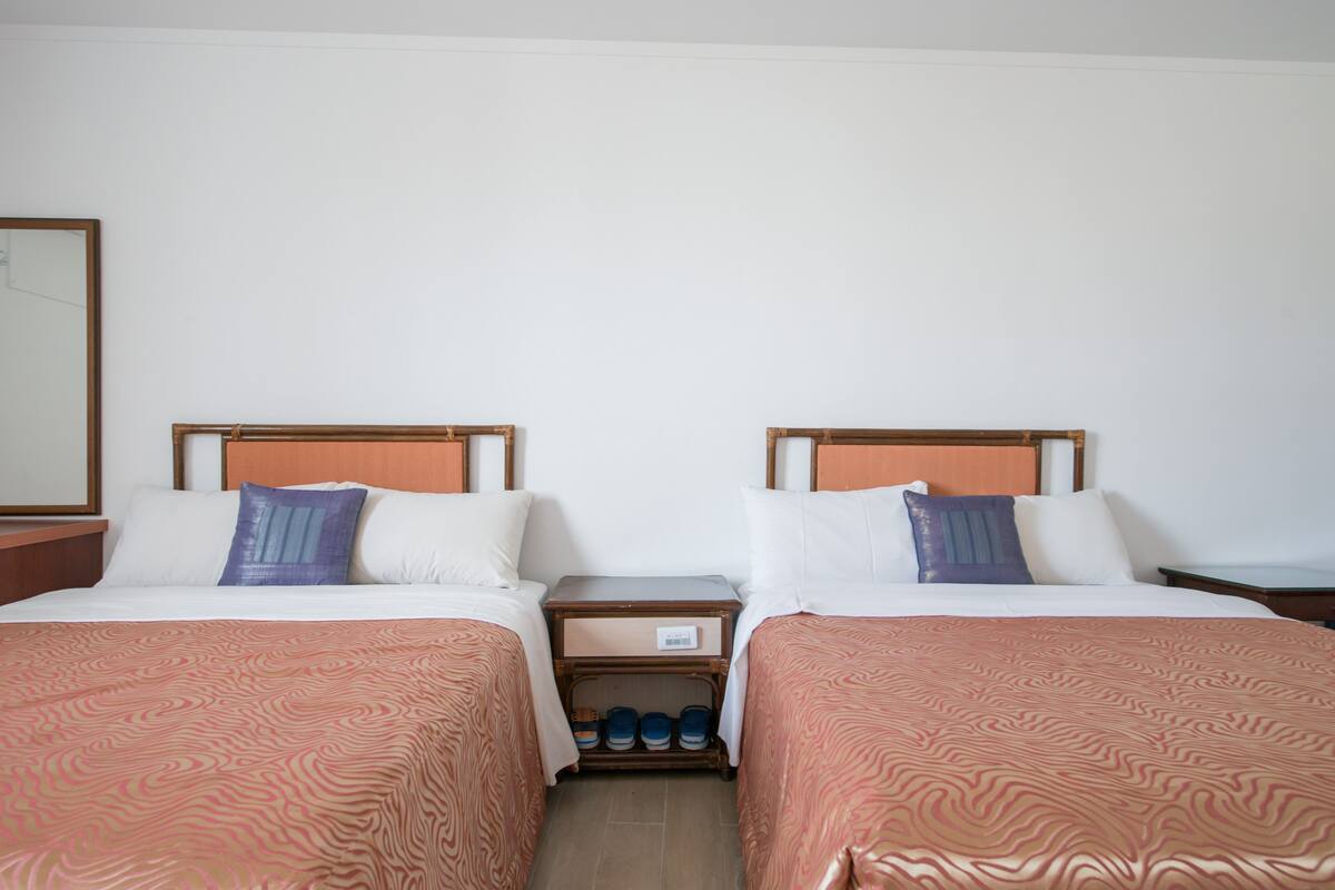 Habitació d'hotel amb dos llits individuals, ambres de fusta, roba de llit blanca i una petita taula de nit de fusta amb prestatge per a sabates.